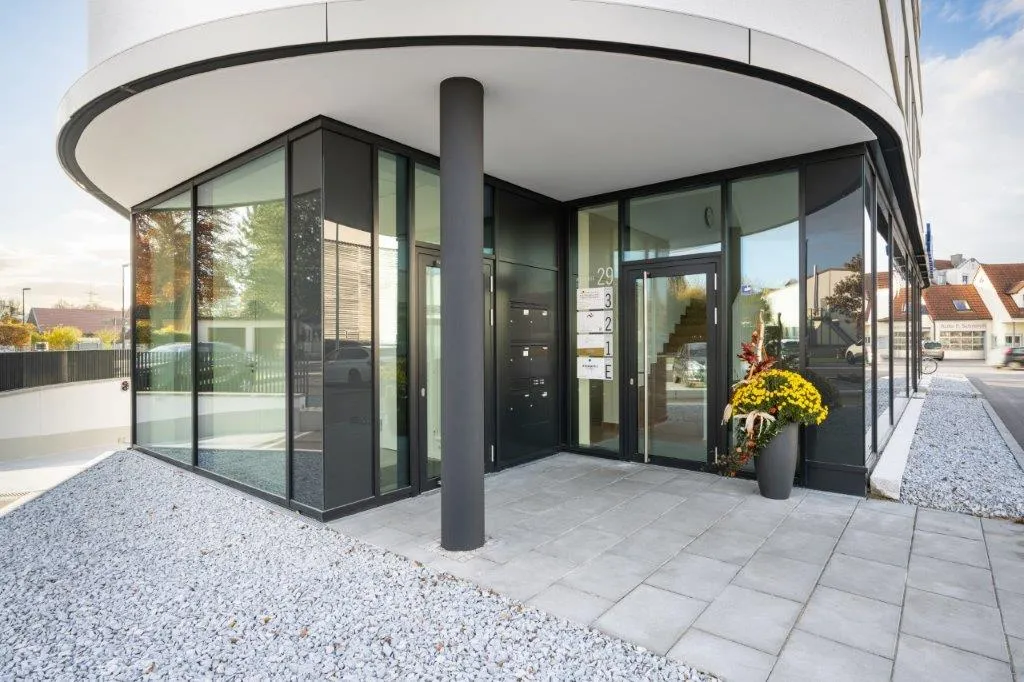 Eingang des runden Neubaus, in dem die Praxis Lenergo liegt, mit Glasfront und Blumen an der Tür